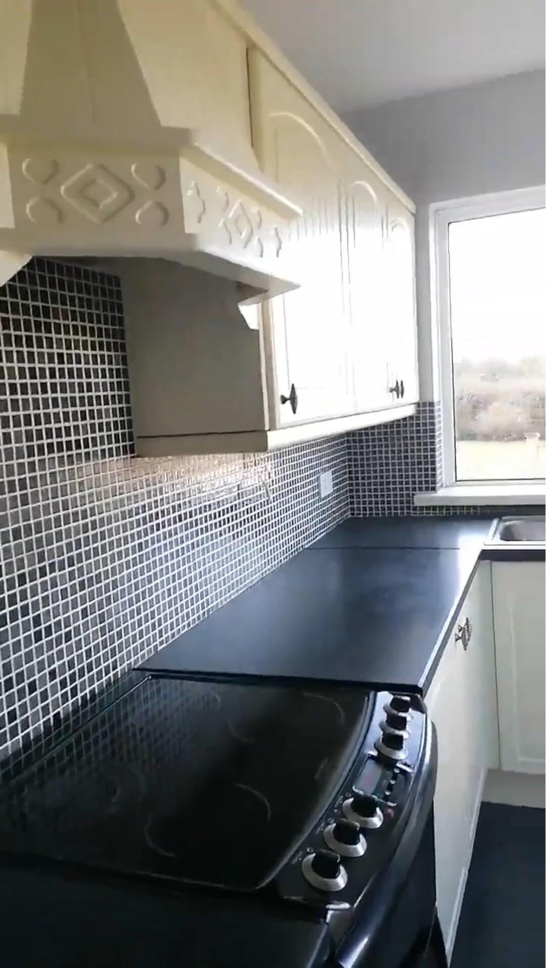 view of a kitchen with a black countertop and cream kitchen units, grey walls and small square black wall tiles